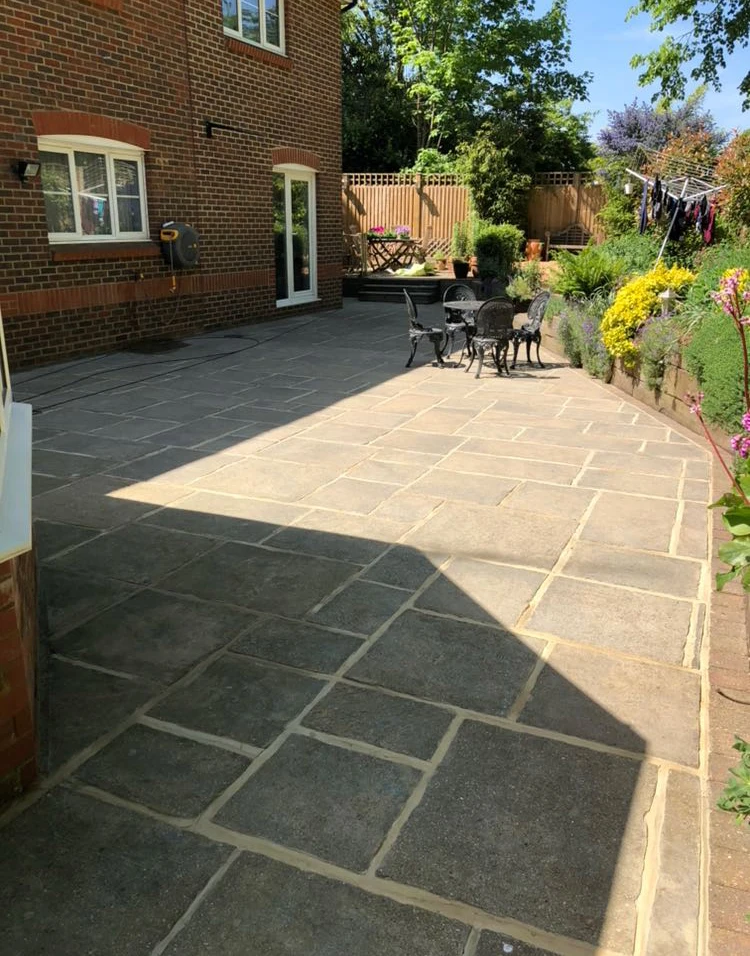 Sunlit stone patio with black metal table and chairs beside brick house and garden with flowers.