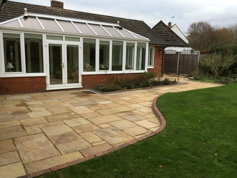 Backyard patio with stone tiles, curved edge lawn, and a glass conservatory attached to a brick house.