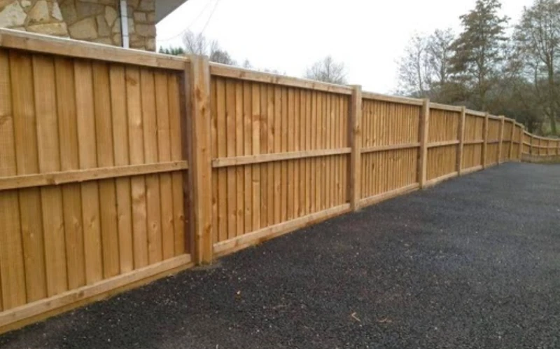 Long wooden fence along a paved driveway with trees and stone building in the background.