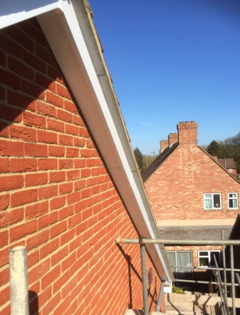 Red brick house wall and roof edge with a metal railing and neighboring houses under a clear blue sky.