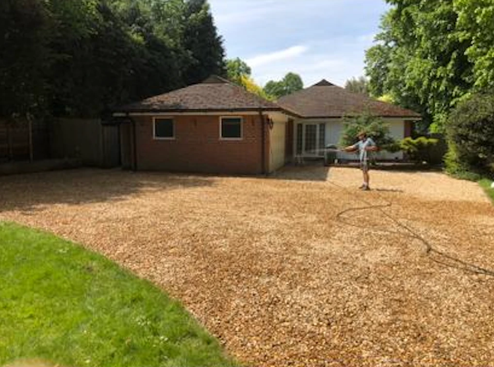 Man standing on a large gravel driveway in front of a brick garage attached to a house.
