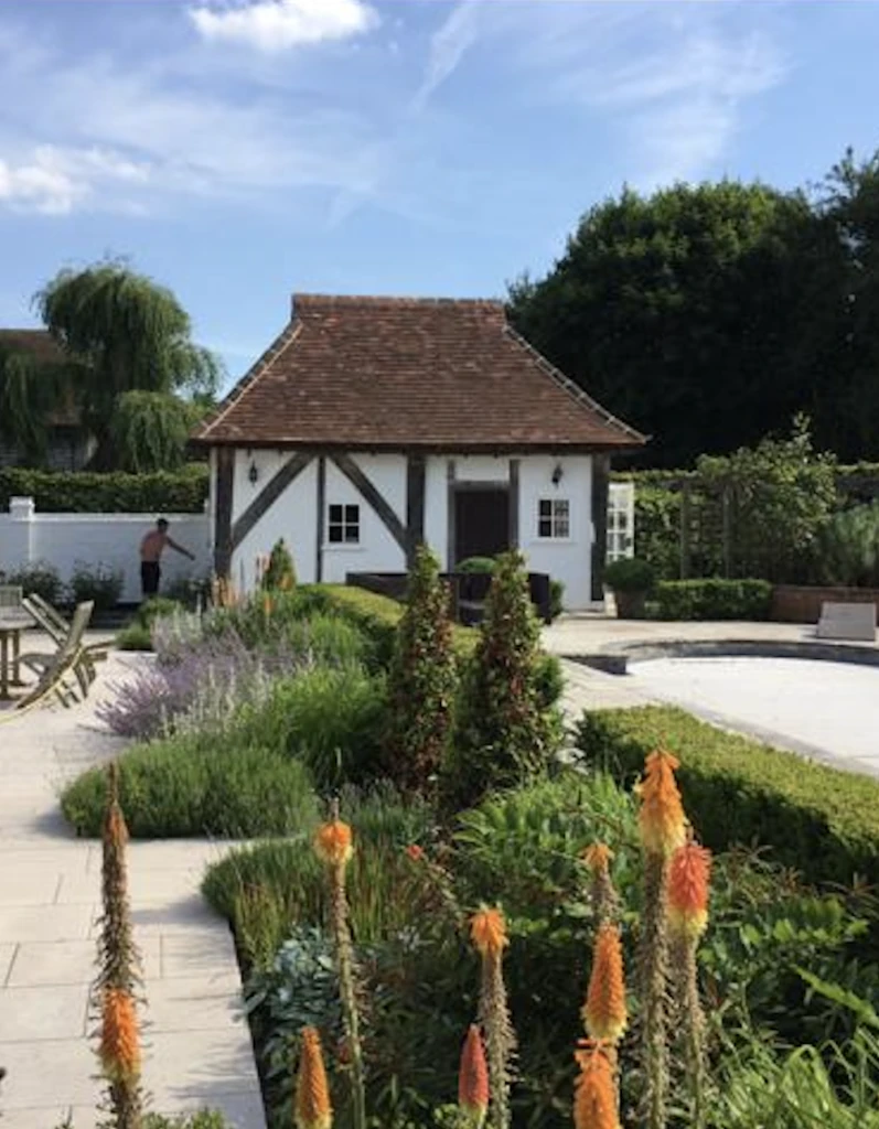 Garden with tall orange flowers and hedges leading to a small white cottage with a brown roof.