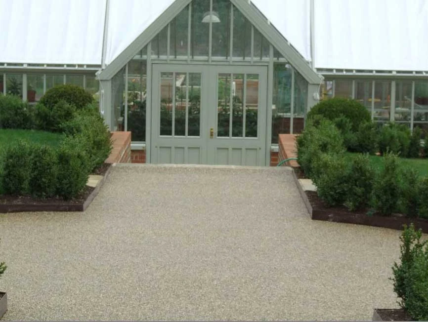 Entrance to a greenhouse with gray framed glass doors, flanked by trimmed green shrubs and brick walls.