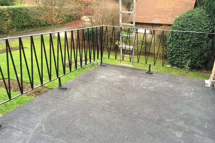 Flat rooftop with black metal railing and a garden view containing grass, shrubs, and trees.