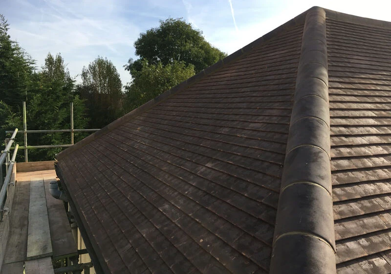 Sloped brown tiled roof corner with ridge tile and scaffolding alongside under a blue sky.